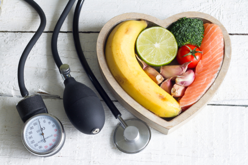 Picture of a blood pressure cuff, stethoscope, and a wooden shaped bowl that has a banana, lime, broccoli, tomato, garlic, lime, and salmon.