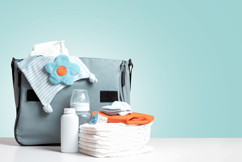 Picture of a baby diaper bag that has baby clothes, a bottle, baby powder, diapers, and a paci sitting outside of the bag on a table.